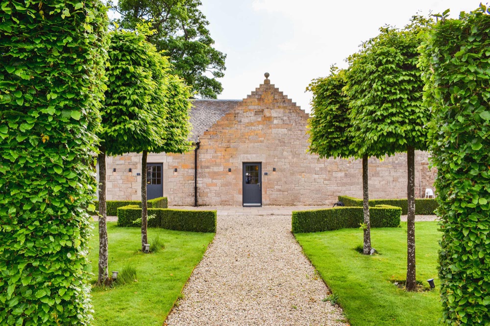 The Stables at Carlowrie: a stone gable and grey door at the end of a gravel path between pleached trees